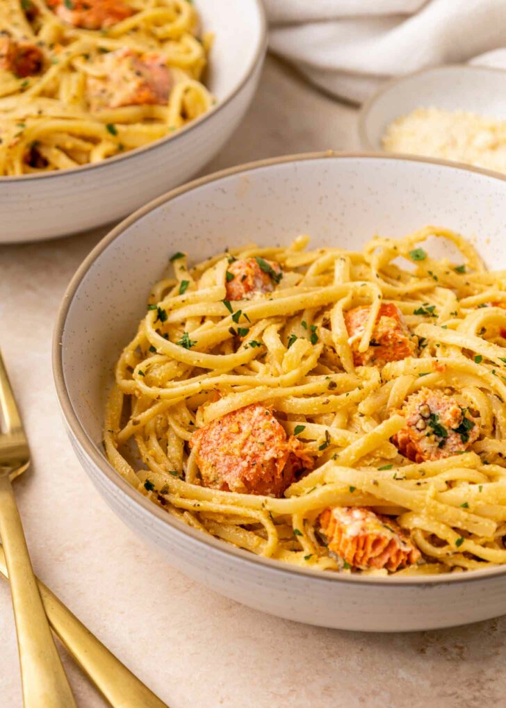 A close-up of a bowl of creamy pasta with chunks of salmon, garnished with chopped herbs. Another bowl and a plate of grated cheese are in the background, alongside gold-colored utensils.