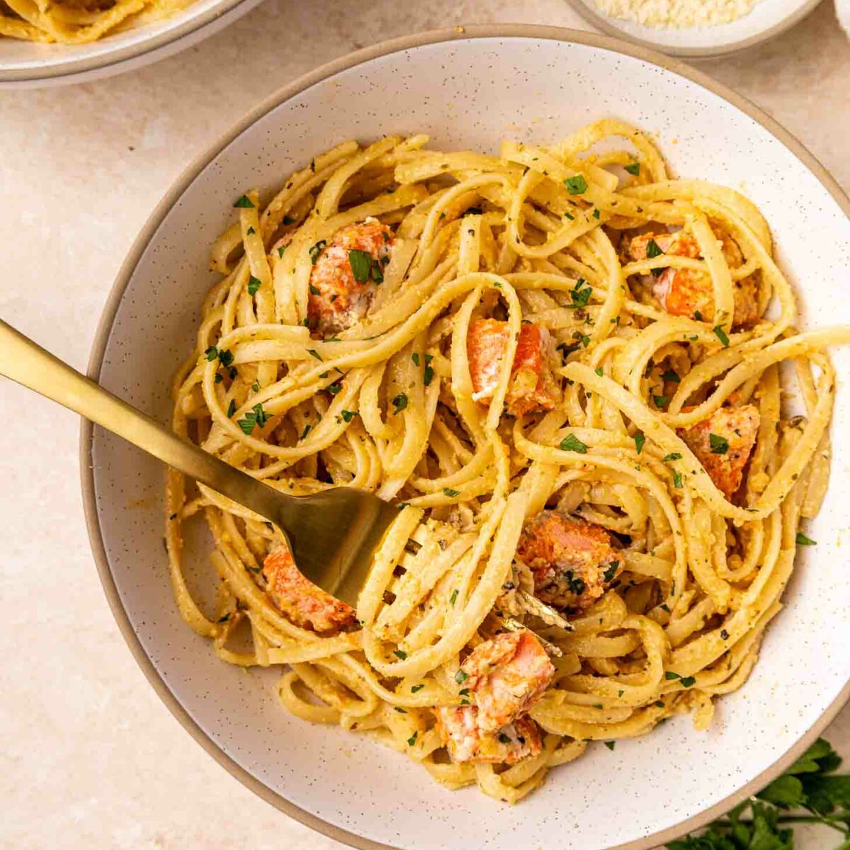A bowl of creamy pasta with chunks of salmon, garnished with chopped parsley. A gold fork is twirled in the pasta, and a small bowl of grated cheese and parsley are nearby on a light-colored surface.