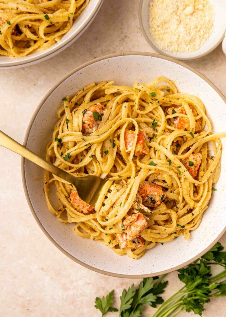 A bowl of creamy pasta with chunks of salmon, garnished with chopped parsley. A gold fork is twirled in the pasta, and a small bowl of grated cheese and parsley are nearby on a light-colored surface.