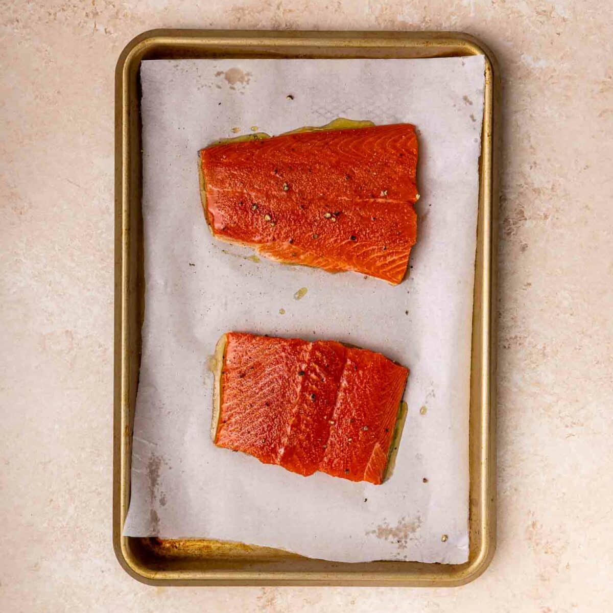 Two raw salmon fillets on a parchment-lined baking sheet, placed on a light-colored countertop. The fillets are seasoned and spaced apart on the tray.
