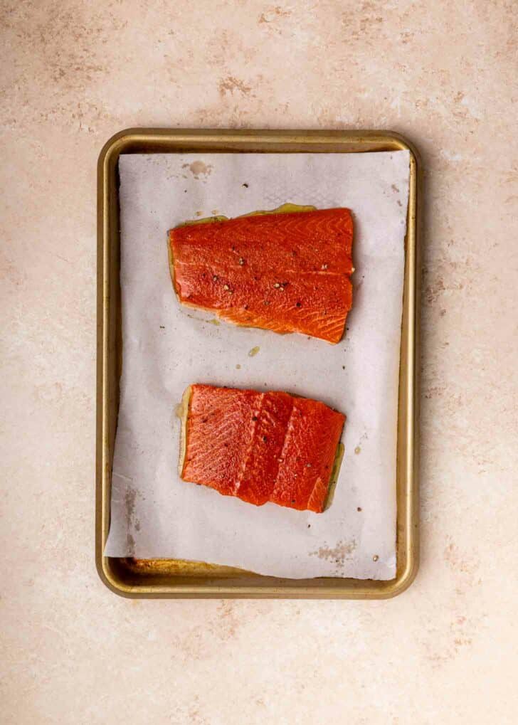 Two raw salmon fillets on a parchment-lined baking sheet, placed on a light-colored countertop. The fillets are seasoned and spaced apart on the tray.