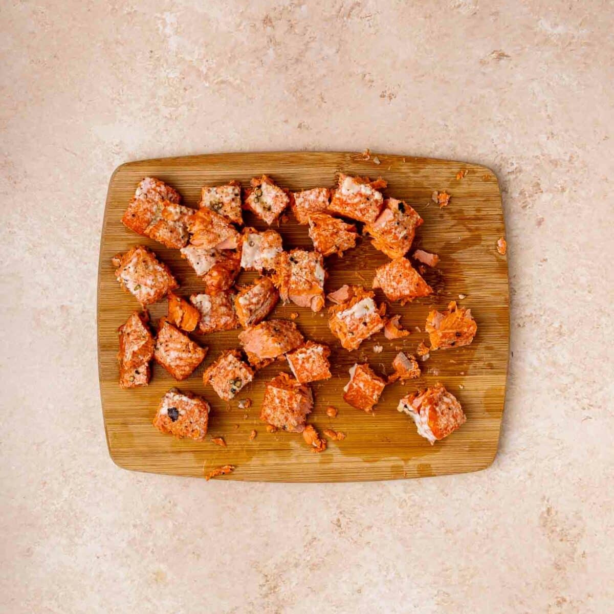 A wooden cutting board with several cubes of cooked salmon scattered on it, set on a light beige countertop.