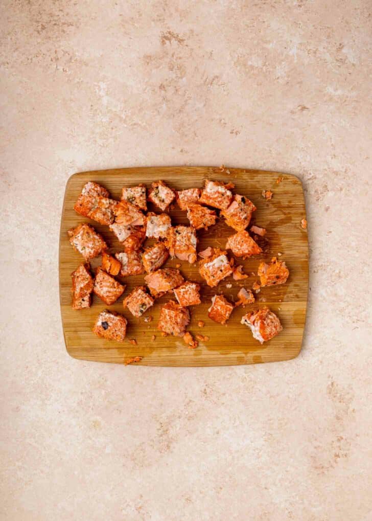 A wooden cutting board with several cubes of cooked salmon scattered on it, set on a light beige countertop.