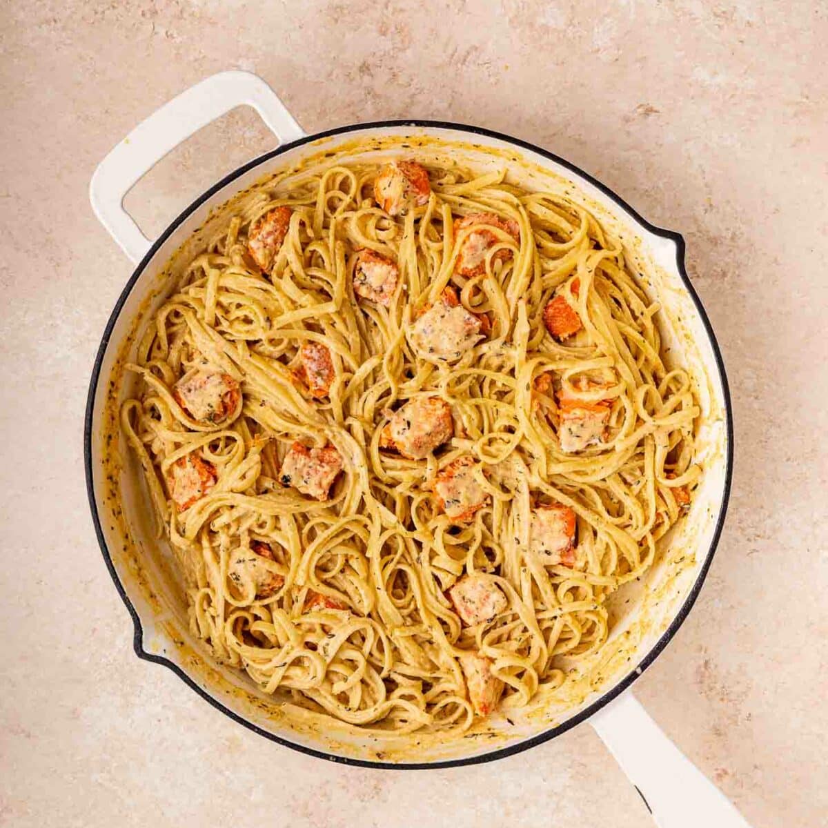 A white skillet filled with creamy pasta and chunks of cooked salmon sits on a light-colored countertop.