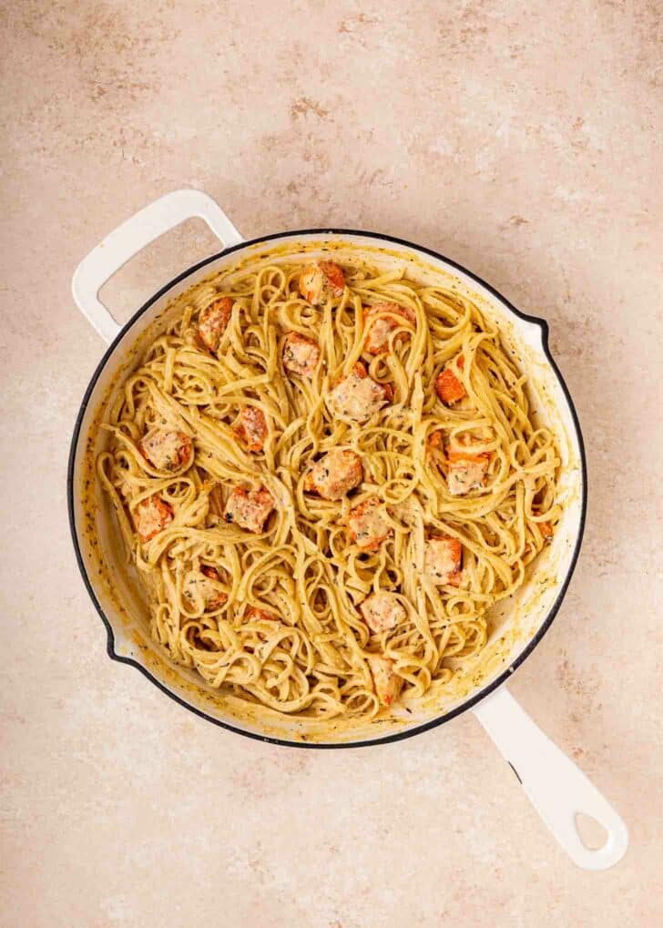 A white skillet filled with creamy pasta and chunks of cooked salmon sits on a light-colored countertop.