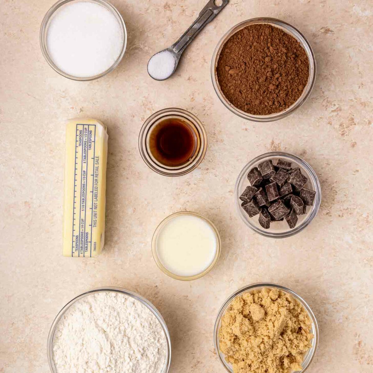 Top-down view of baking ingredients in bowls: sugar, cocoa powder, chocolate chunks, flour, brown sugar, a stick of butter, vanilla extract, salt in a measuring spoon, and a small bowl of liquid on a light surface.