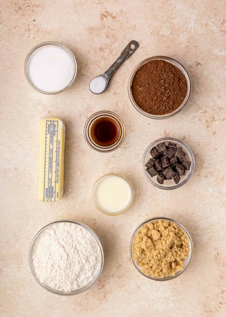 Top-down view of baking ingredients in bowls: sugar, cocoa powder, chocolate chunks, flour, brown sugar, a stick of butter, vanilla extract, salt in a measuring spoon, and a small bowl of liquid on a light surface.