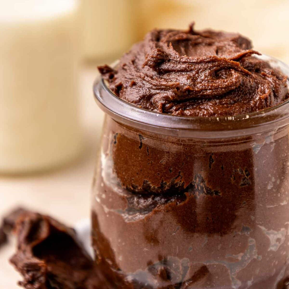 A small glass jar filled with rich, creamy brownie batter sits on a white saucer. A spoon with chocolate mousse rests beside the jar. The background is softly blurred with light colors.