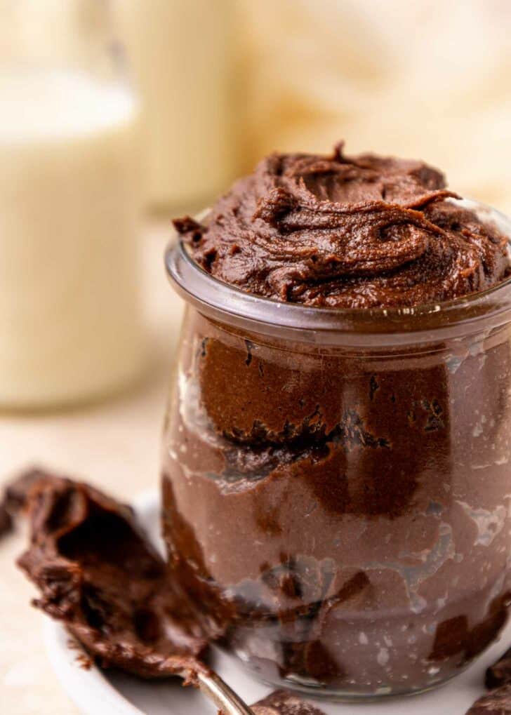 A small glass jar filled with rich, creamy brownie batter sits on a white saucer. A spoon with chocolate mousse rests beside the jar. The background is softly blurred with light colors.