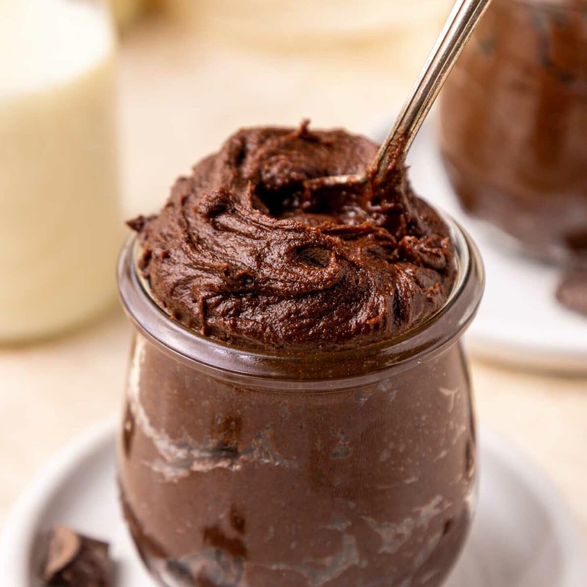 A glass jar filled with rich, creamy brownie batter, with a spoon scooping into the top. Chocolate chunks are scattered on the white plate beneath the jar, and a blurred milk bottle is in the background.