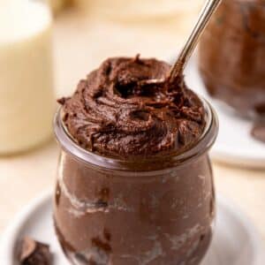 A glass jar filled with rich, creamy brownie batter, with a spoon scooping into the top. Chocolate chunks are scattered on the white plate beneath the jar, and a blurred milk bottle is in the background.