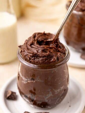 A glass jar filled with rich, creamy brownie batter, with a spoon scooping into the top. Chocolate chunks are scattered on the white plate beneath the jar, and a blurred milk bottle is in the background.