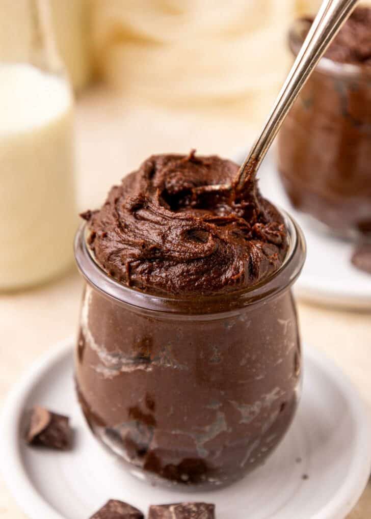A glass jar filled with rich, creamy brownie batter, with a spoon scooping into the top. Chocolate chunks are scattered on the white plate beneath the jar, and a blurred milk bottle is in the background.