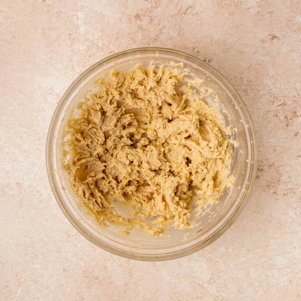 A glass bowl filled with creamed brownie batter sits on a light beige, textured surface. The dough has a pale, slightly crumbly appearance and is evenly distributed within the bowl.