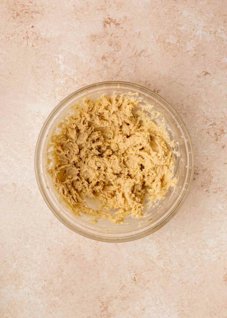 A glass bowl filled with creamed brownie batter sits on a light beige, textured surface. The dough has a pale, slightly crumbly appearance and is evenly distributed within the bowl.