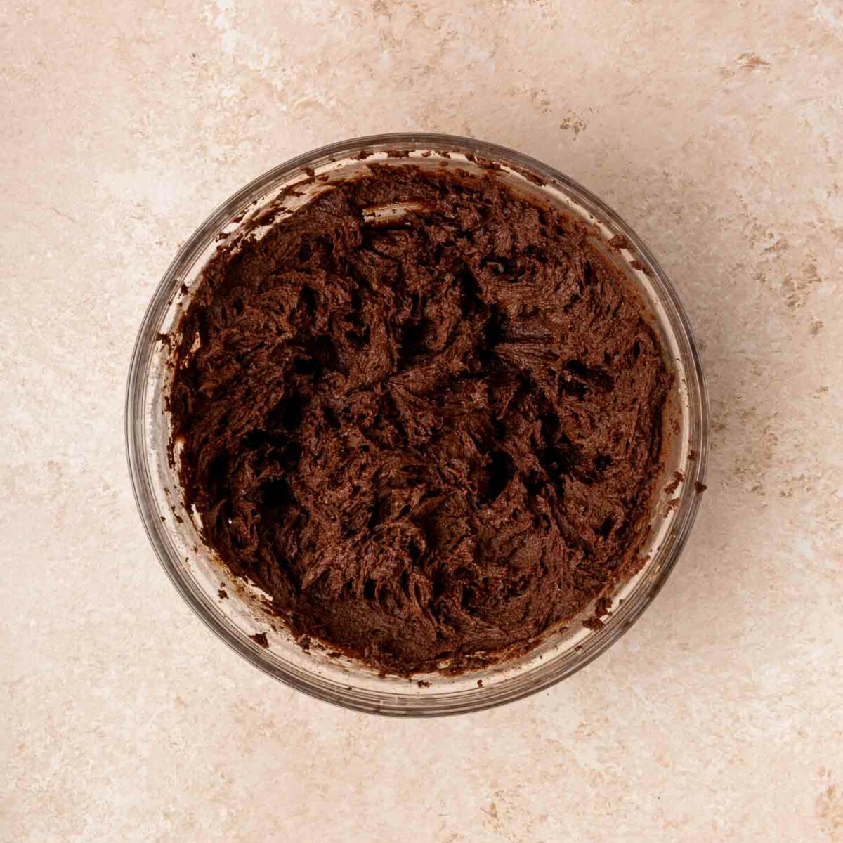 A glass bowl filled with creamy brownie batter sits on a light beige countertop.