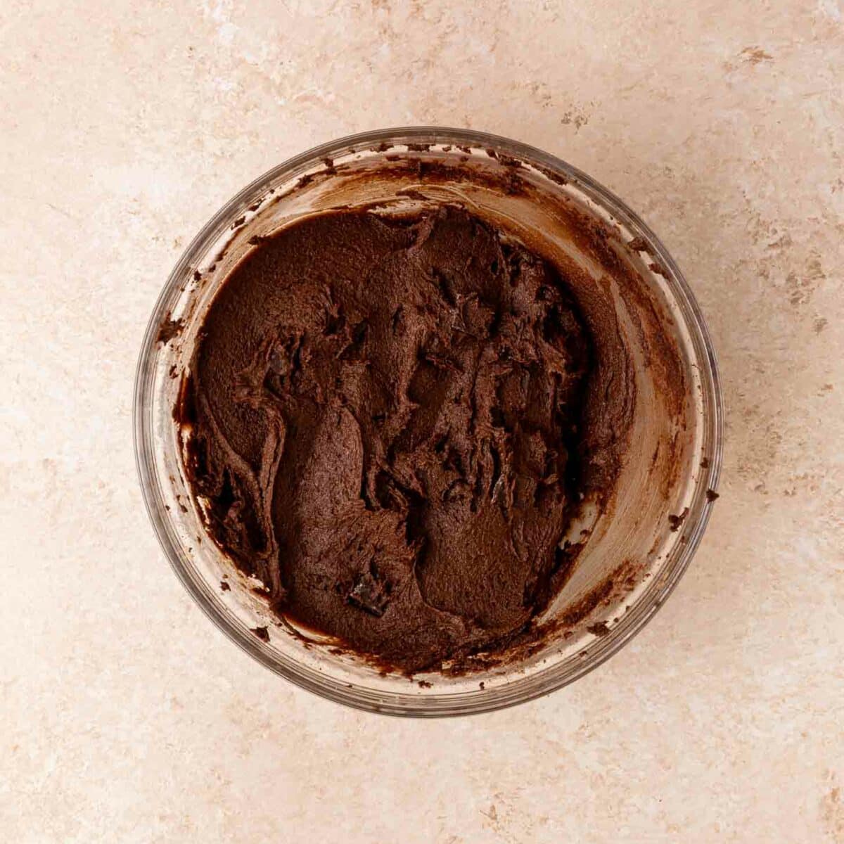 A glass bowl filled with thick, creamy brownie batter sits on a light beige countertop. The frosting is spread unevenly with visible swirls and chocolate chunks.