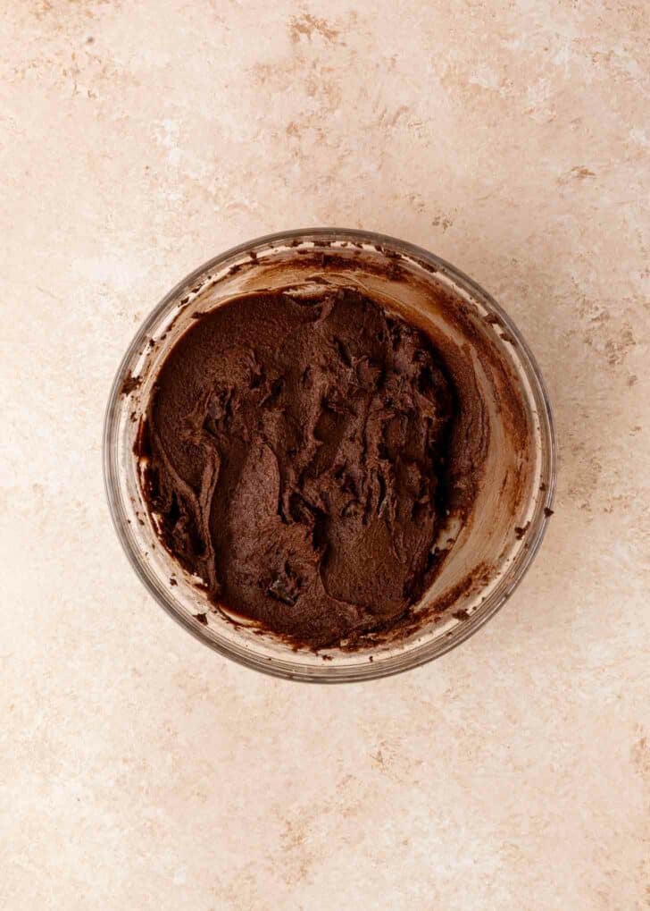 A glass bowl filled with thick, creamy brownie batter sits on a light beige countertop. The frosting is spread unevenly with visible swirls and chocolate chunks.