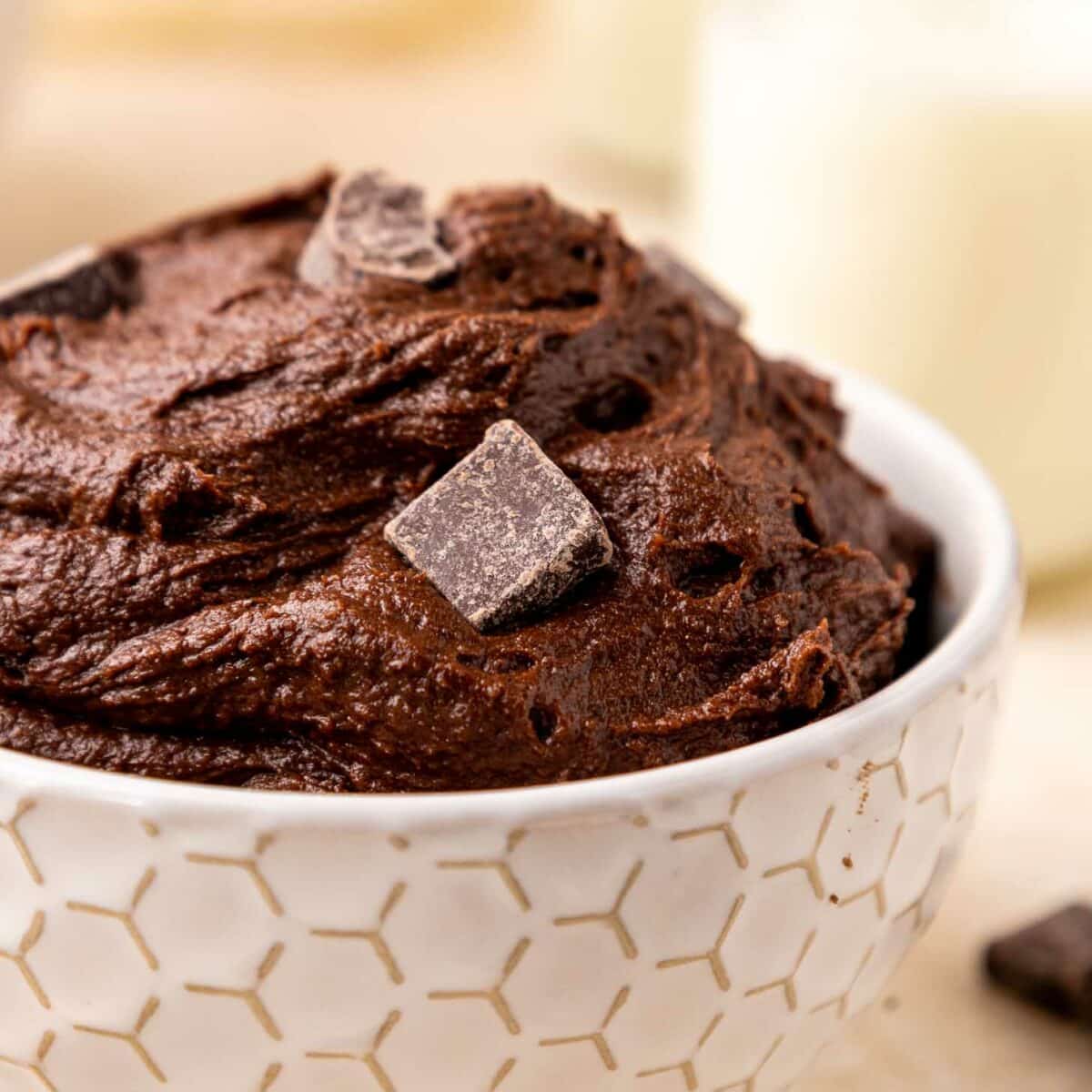 A white bowl with a geometric pattern filled with rich brownie batter, topped with a few chunks of dark chocolate.