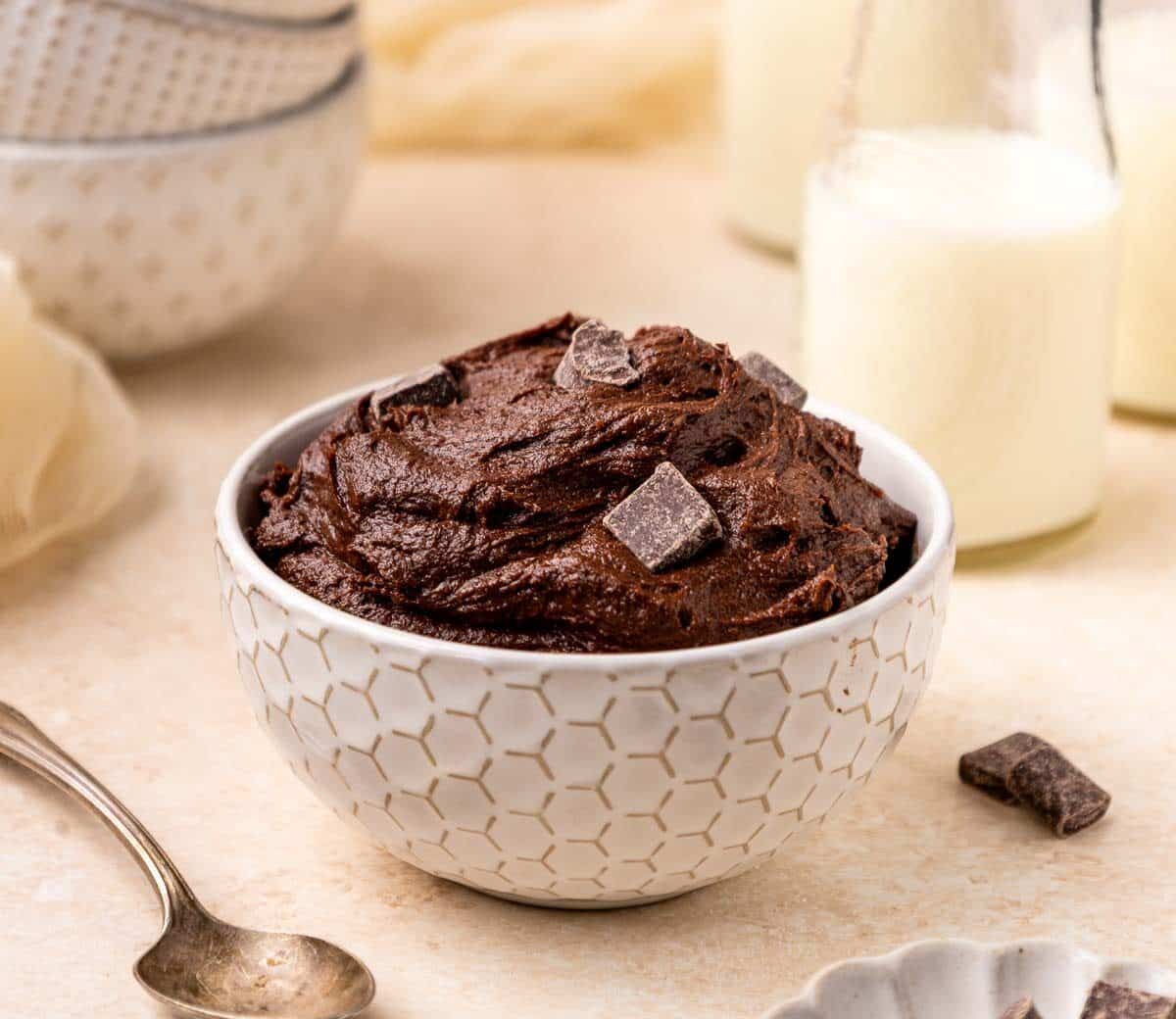 A white patterned bowl filled with chocolate brownie batter dough topped with chocolate chunks, placed on a light surface with a spoon, glass bottles of milk, and stacked bowls in the background.