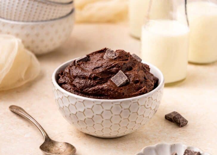 A white patterned bowl filled with chocolate brownie batter dough topped with chocolate chunks, placed on a light surface with a spoon, glass bottles of milk, and stacked bowls in the background.