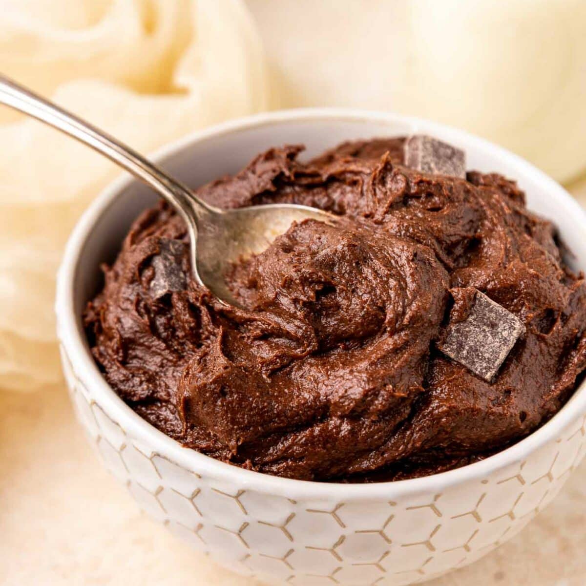 A white textured bowl filled with rich brownie batter, topped with chocolate chunks, and a spoon partially dipped into the dough. The background includes a light cloth and a glass of milk.