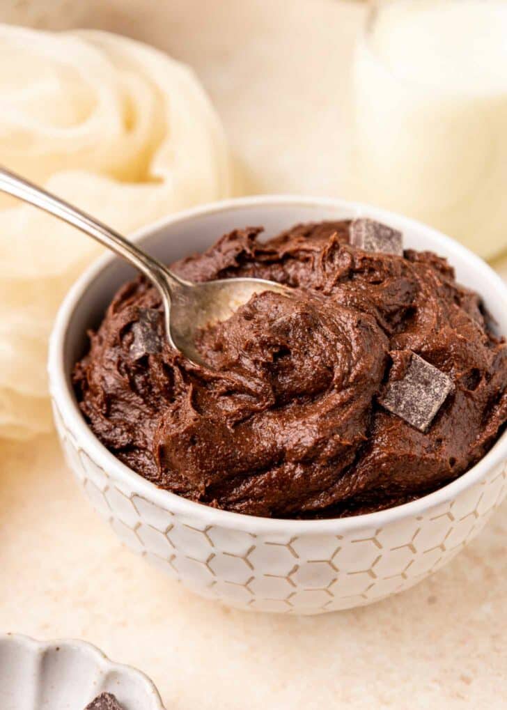 A white textured bowl filled with rich brownie batter, topped with chocolate chunks, and a spoon partially dipped into the dough. The background includes a light cloth and a glass of milk.