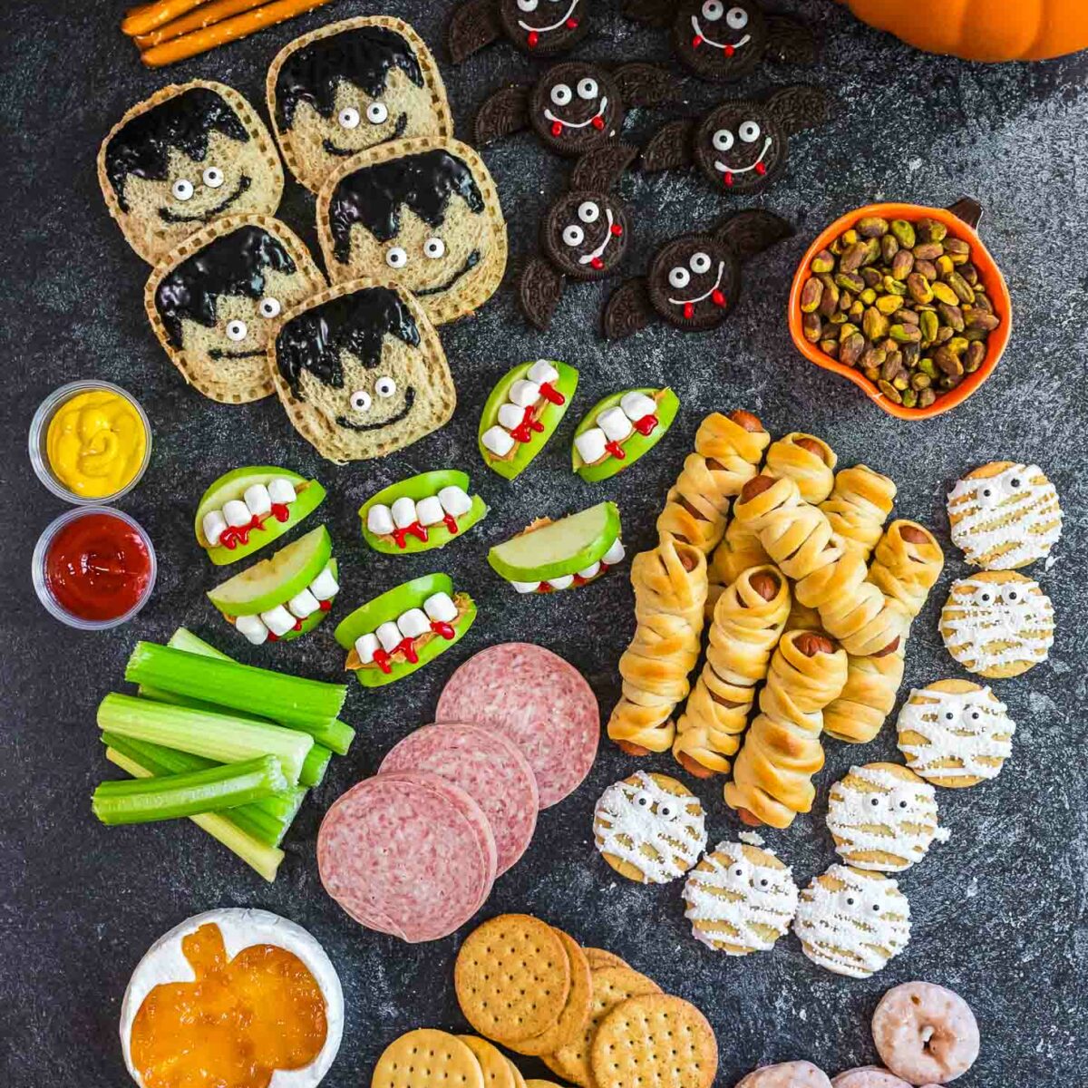 A Halloween-themed snack platter with treats shaped like mummies, monsters, and vampires, apple slices with candy teeth, crackers, pretzels, dipping sauces, pistachios, cookies, and donuts, surrounded by two pumpkins.
