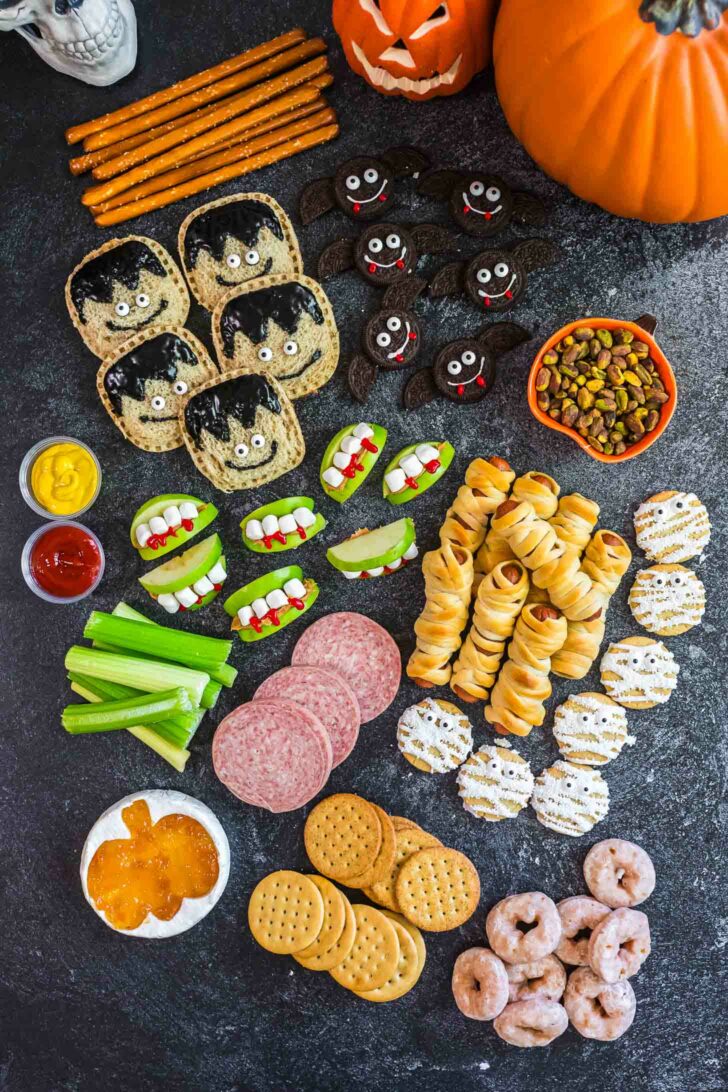 A Halloween-themed snack platter with treats shaped like mummies, monsters, and vampires, apple slices with candy teeth, crackers, pretzels, dipping sauces, pistachios, cookies, and donuts, surrounded by two pumpkins.