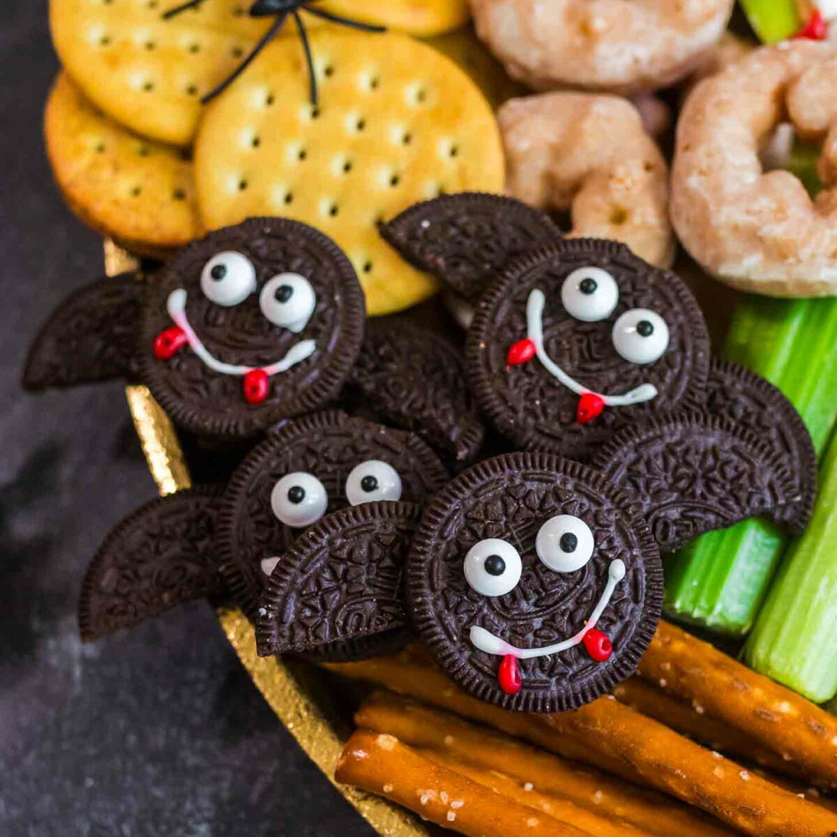 A festive snack platter features bat-shaped cookies made from chocolate sandwich cookies with candy eyes and icing smiles, surrounded by crackers, donuts, celery, and pretzel sticks. A fake spider sits on the crackers.
