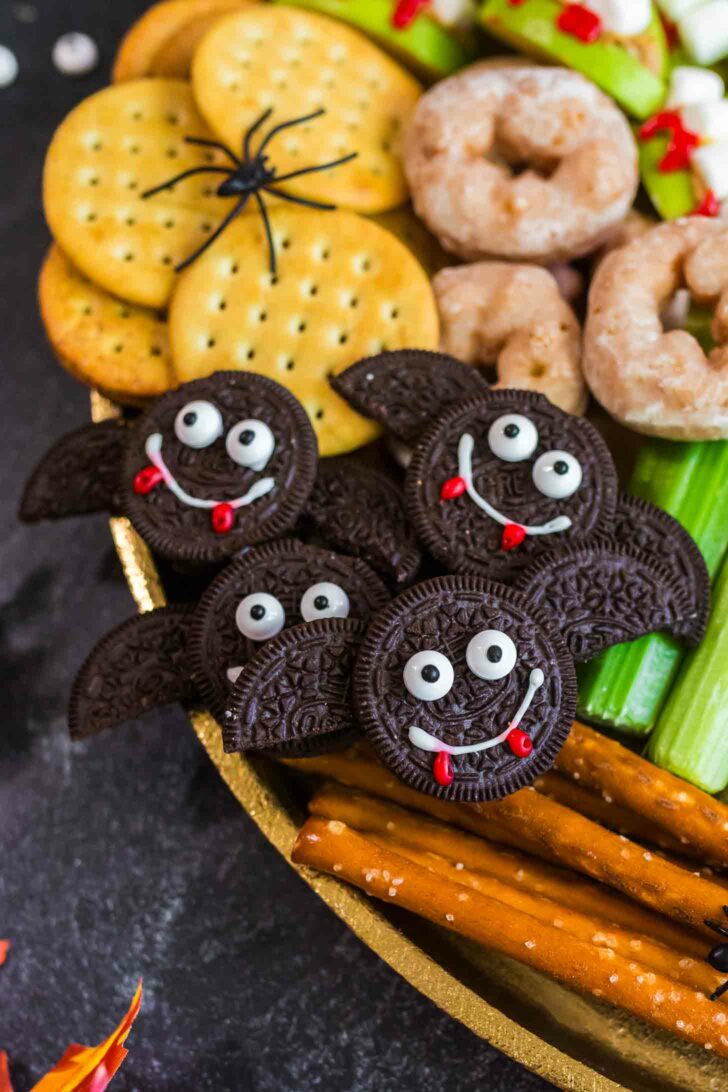 A festive snack platter features bat-shaped cookies made from chocolate sandwich cookies with candy eyes and icing smiles, surrounded by crackers, donuts, celery, and pretzel sticks. A fake spider sits on the crackers.
