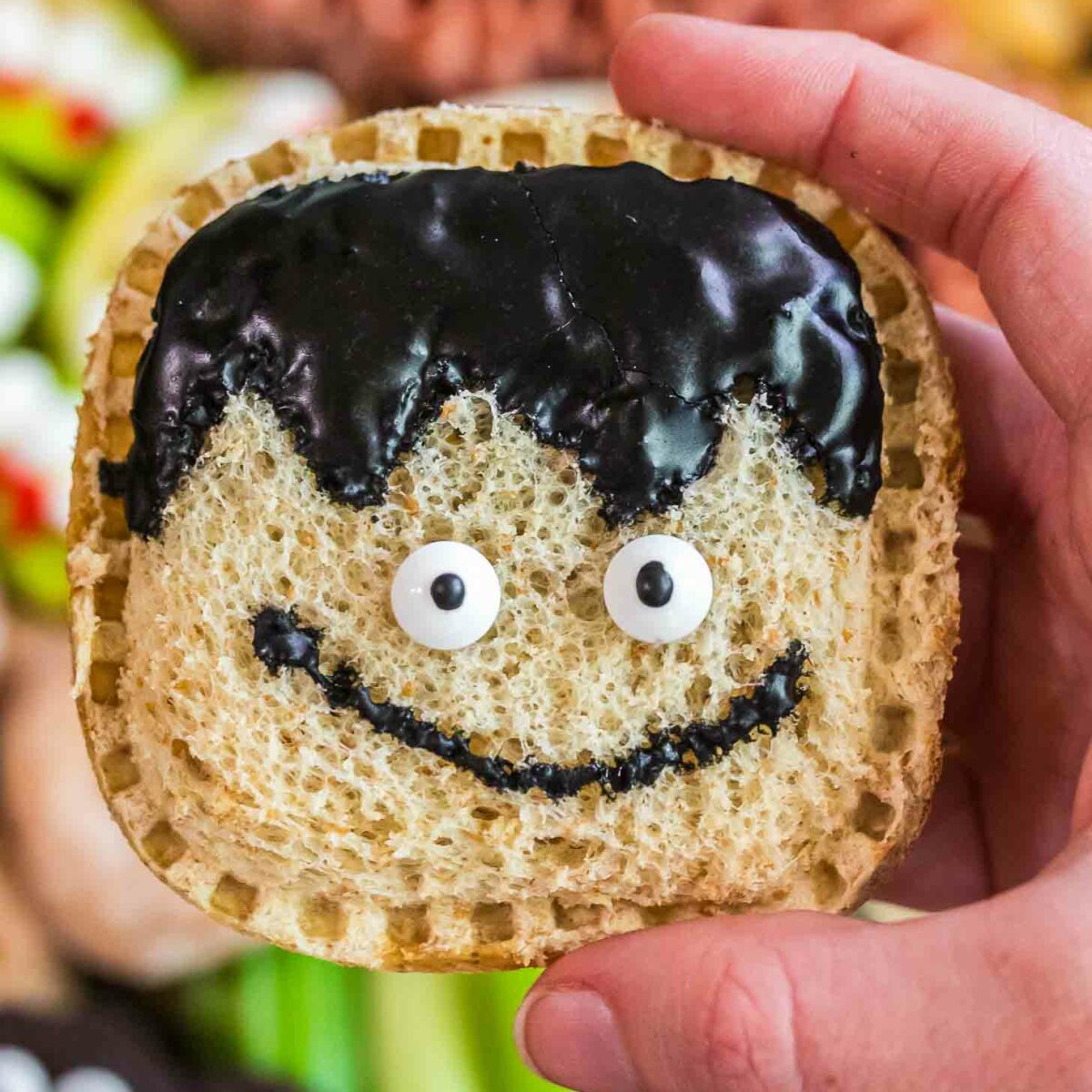 A hand holds a square sandwich with a whimsical face made from candy eyes and black icing shaped like hair and a smile, resembling a cartoon character. The background features assorted snacks.