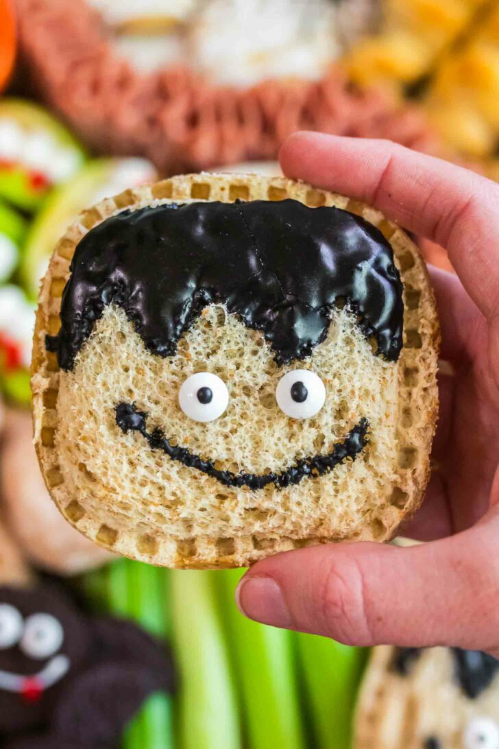 A hand holds a square sandwich with a whimsical face made from candy eyes and black icing shaped like hair and a smile, resembling a cartoon character. The background features assorted snacks.