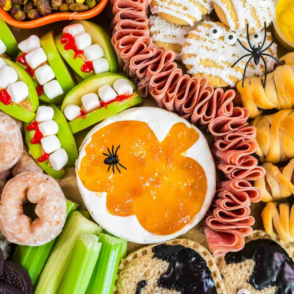 A Halloween-themed snack platter with apple and marshmallow “monster mouths,” cookies decorated like mummies, celery sticks, donut holes, crackers with spooky faces, pistachios, cured meat, and cheese topped with orange jelly and fake spiders.