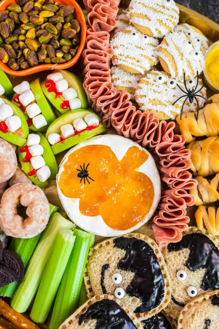 A Halloween-themed snack platter with apple and marshmallow “monster mouths,” cookies decorated like mummies, celery sticks, donut holes, crackers with spooky faces, pistachios, cured meat, and cheese topped with orange jelly and fake spiders.