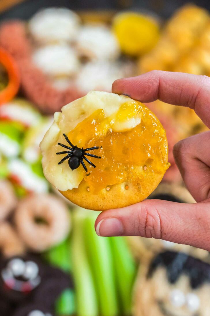 A hand holds a round cracker topped with cheese, orange jam, and a small plastic black spider, with a colorful snack platter blurred in the background.