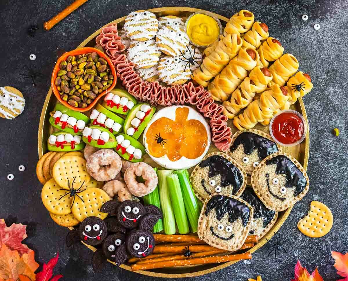 A round platter with Halloween-themed snacks: monster sandwiches, mummy-wrapped treats, apple slices with marshmallow teeth, cookies, cookies with eyes, crackers, meats, and dip shaped like a pumpkin. Autumn leaves border the scene.