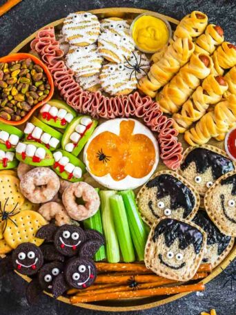 A round platter with Halloween-themed snacks: monster sandwiches, mummy-wrapped treats, apple slices with marshmallow teeth, cookies, cookies with eyes, crackers, meats, and dip shaped like a pumpkin. Autumn leaves border the scene.