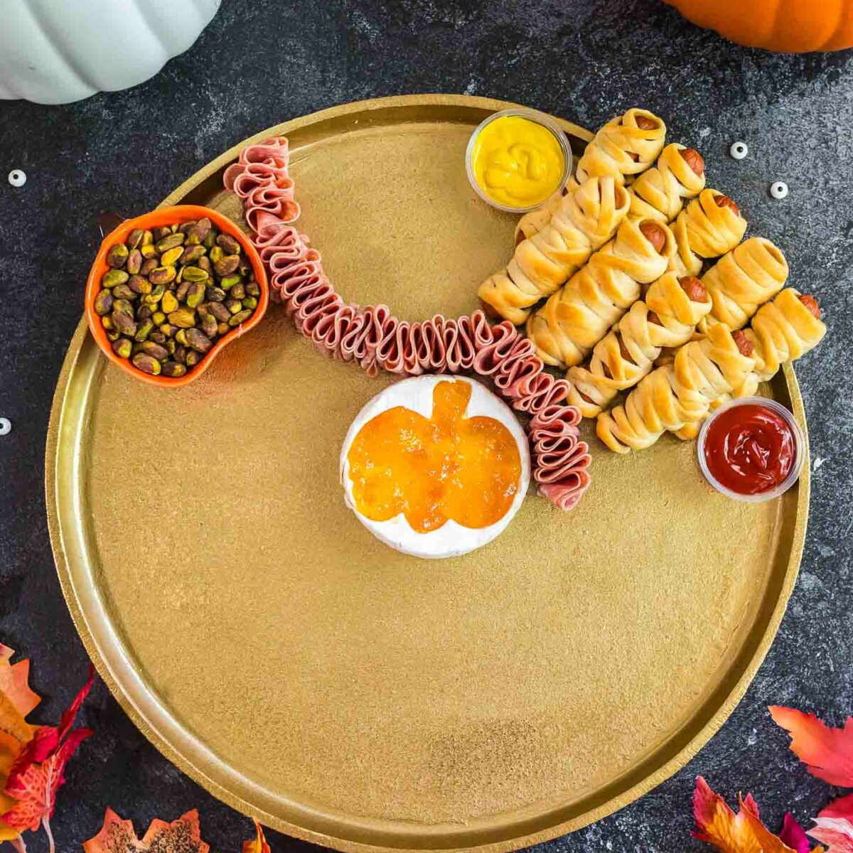 A gold platter with Halloween-themed snacks: mini mummy hot dogs, a line of folded lunchmeat, a pumpkin-shaped bowl of orange dip, mustard, ketchup, and a bowl of nuts, surrounded by pumpkins and autumn leaves.