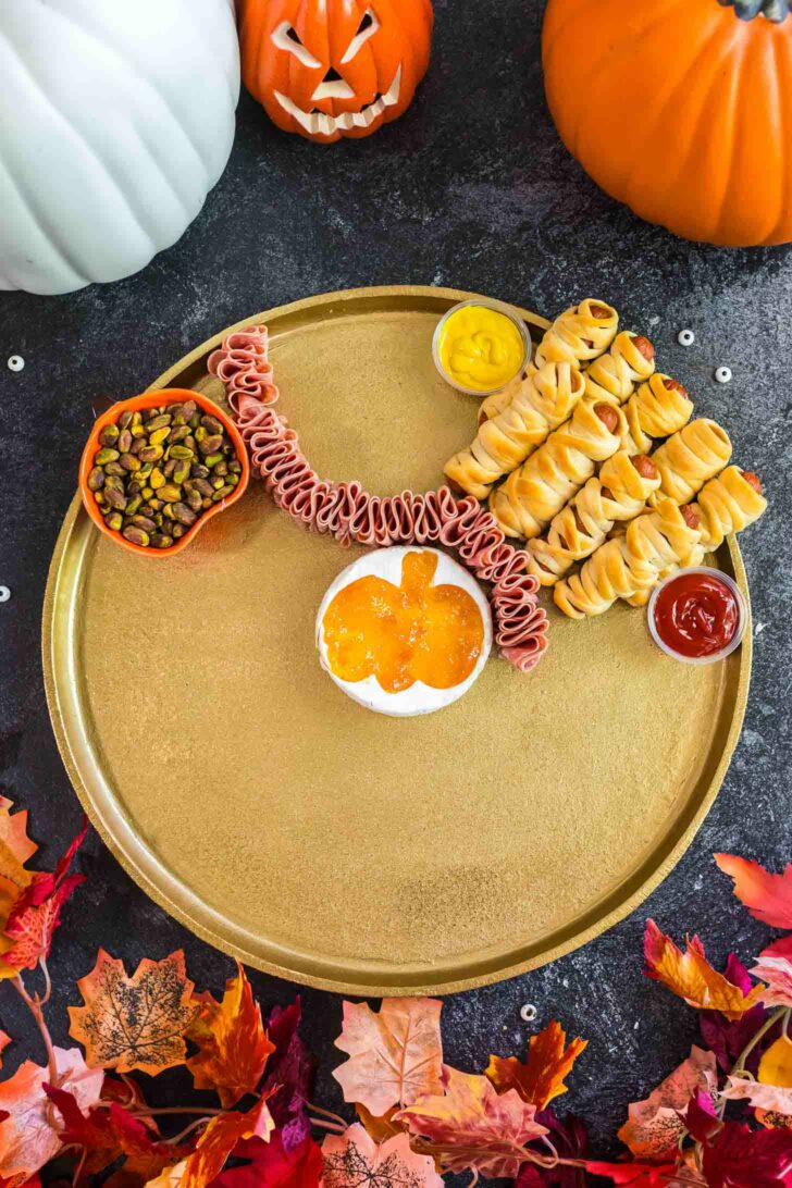 A gold platter with Halloween-themed snacks: mini mummy hot dogs, a line of folded lunchmeat, a pumpkin-shaped bowl of orange dip, mustard, ketchup, and a bowl of nuts, surrounded by pumpkins and autumn leaves.