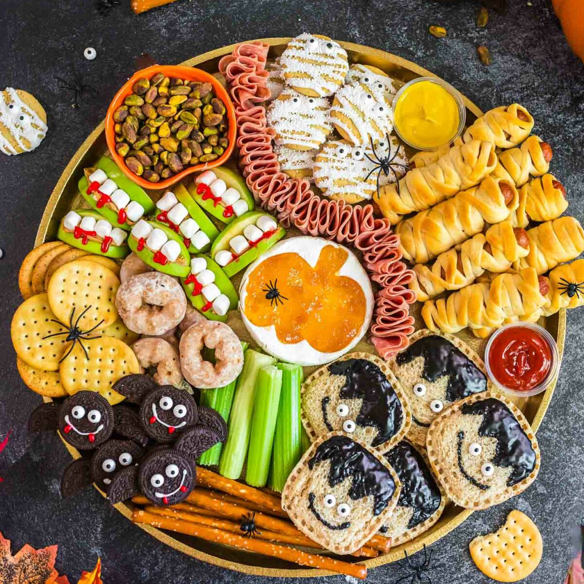 A festive Halloween snack platter with crackers, cookies, celery, donuts, mummy-shaped pastries, apple bites, meats, dips, and spider decorations, surrounded by pumpkins and autumn leaves on a dark surface.