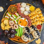 A festive Halloween snack platter with crackers, cookies, celery, donuts, mummy-shaped pastries, apple bites, meats, dips, and spider decorations, surrounded by pumpkins and autumn leaves on a dark surface.