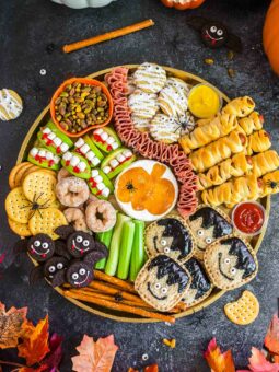 A festive Halloween snack platter with crackers, cookies, celery, donuts, mummy-shaped pastries, apple bites, meats, dips, and spider decorations, surrounded by pumpkins and autumn leaves on a dark surface.