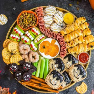 A festive Halloween snack platter with crackers, cookies, celery, donuts, mummy-shaped pastries, apple bites, meats, dips, and spider decorations, surrounded by pumpkins and autumn leaves on a dark surface.