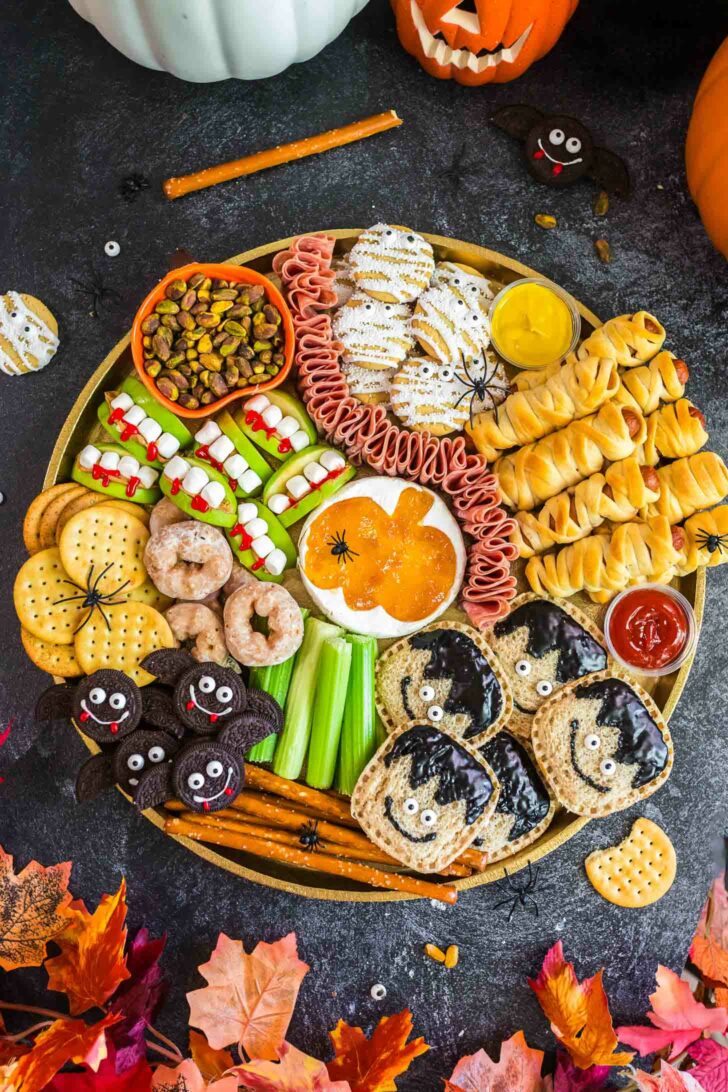 A festive Halloween snack platter with crackers, cookies, celery, donuts, mummy-shaped pastries, apple bites, meats, dips, and spider decorations, surrounded by pumpkins and autumn leaves on a dark surface.