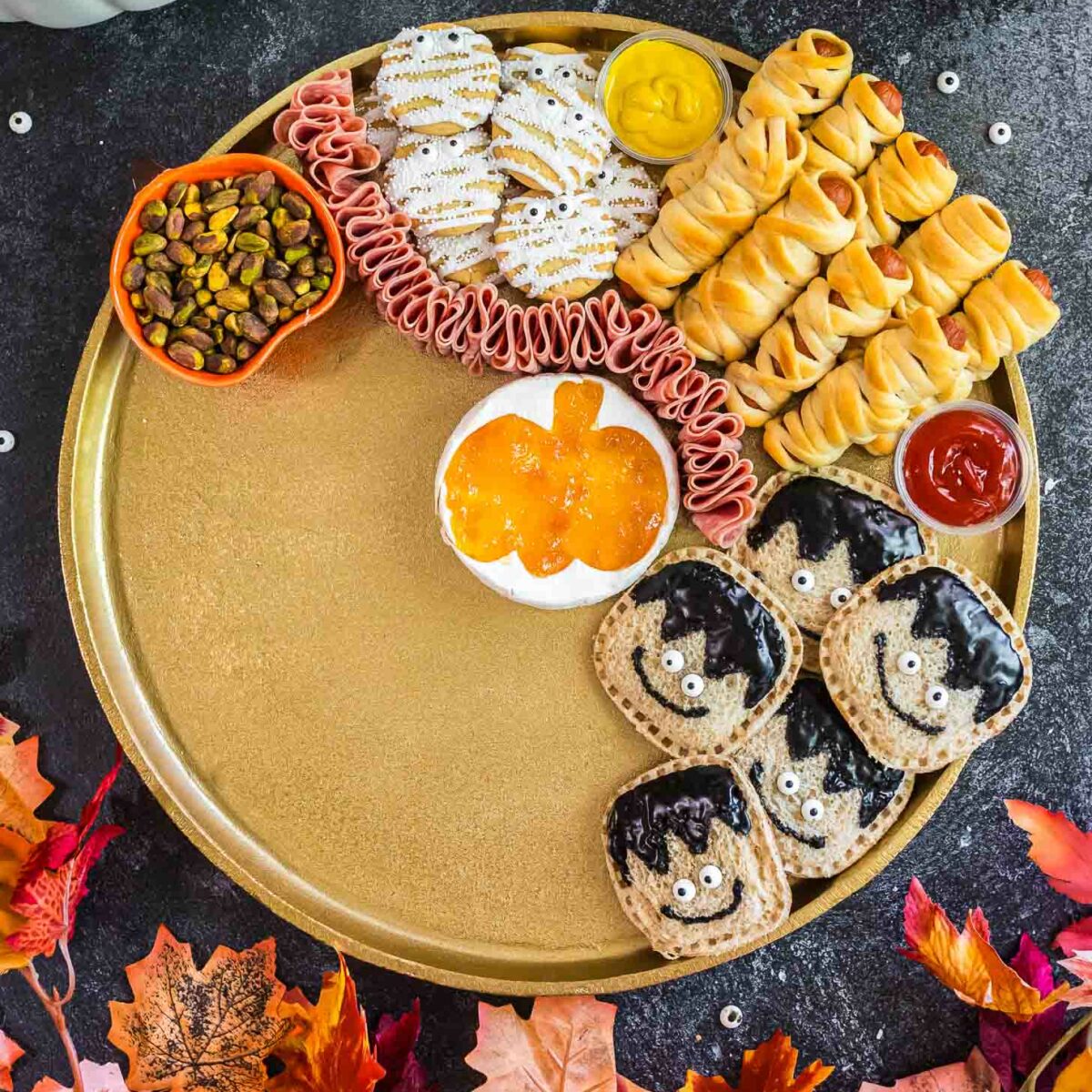 A festive platter with Halloween-themed snacks: cookies with white icing, rolled deli meat, mini hot dogs wrapped like mummies, sauces, pistachios, jam, and bread rounds decorated as bats. Autumn leaves and pumpkins surround the plate.