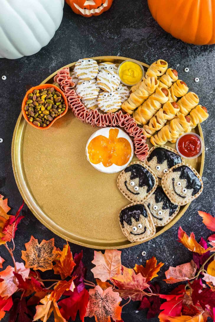 A festive platter with Halloween-themed snacks: cookies with white icing, rolled deli meat, mini hot dogs wrapped like mummies, sauces, pistachios, jam, and bread rounds decorated as bats. Autumn leaves and pumpkins surround the plate.