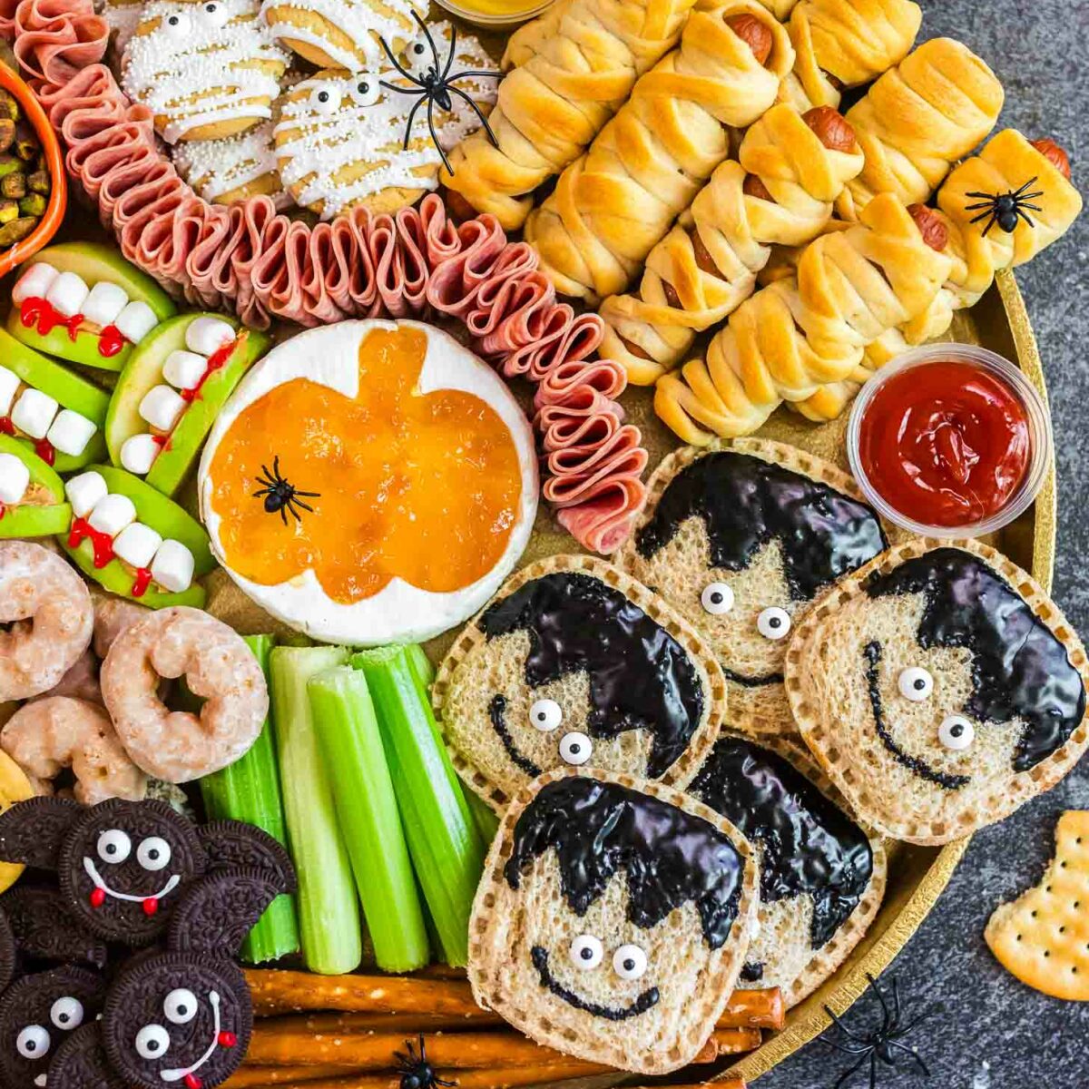 A round platter with Halloween-themed snacks, including mummy-wrapped sausages, ghost cookies, monster sandwiches, veggie sticks resembling monsters, bat-shaped cookies, pretzels, candy eyes, and dips for a festive party.