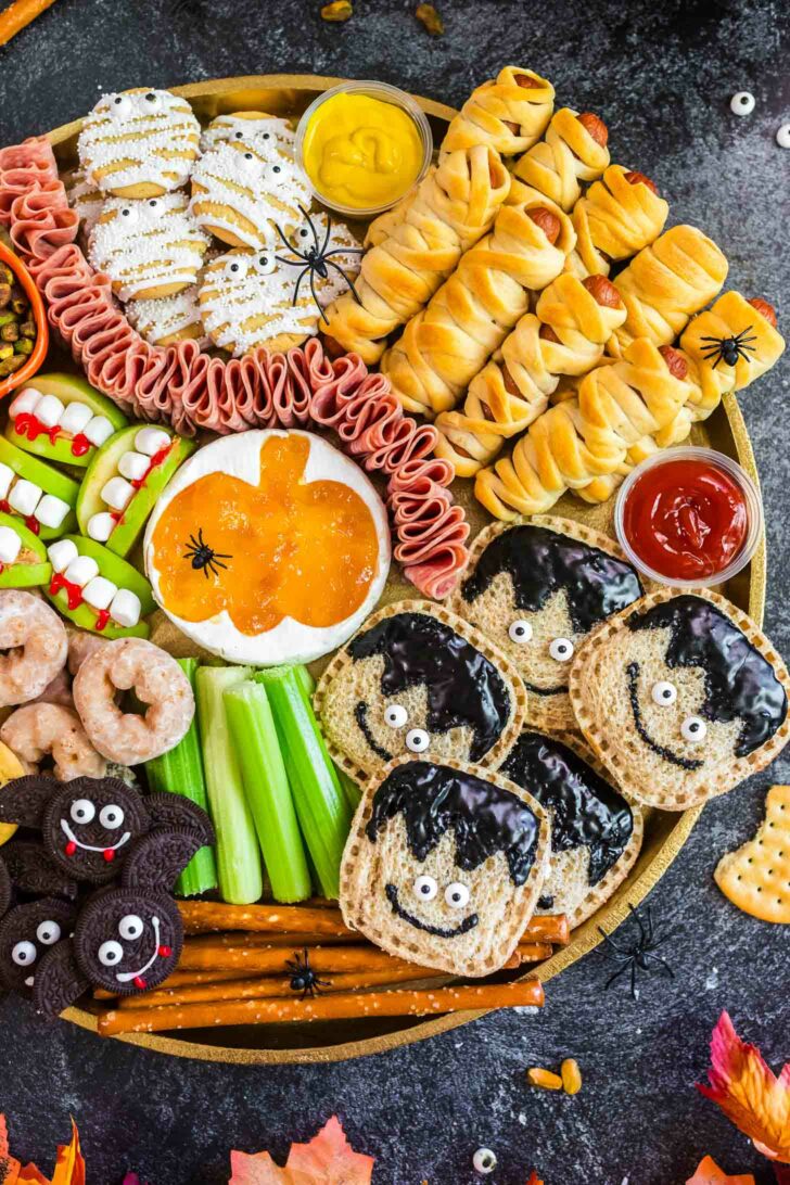 A round platter with Halloween-themed snacks, including mummy-wrapped sausages, ghost cookies, monster sandwiches, veggie sticks resembling monsters, bat-shaped cookies, pretzels, candy eyes, and dips for a festive party.