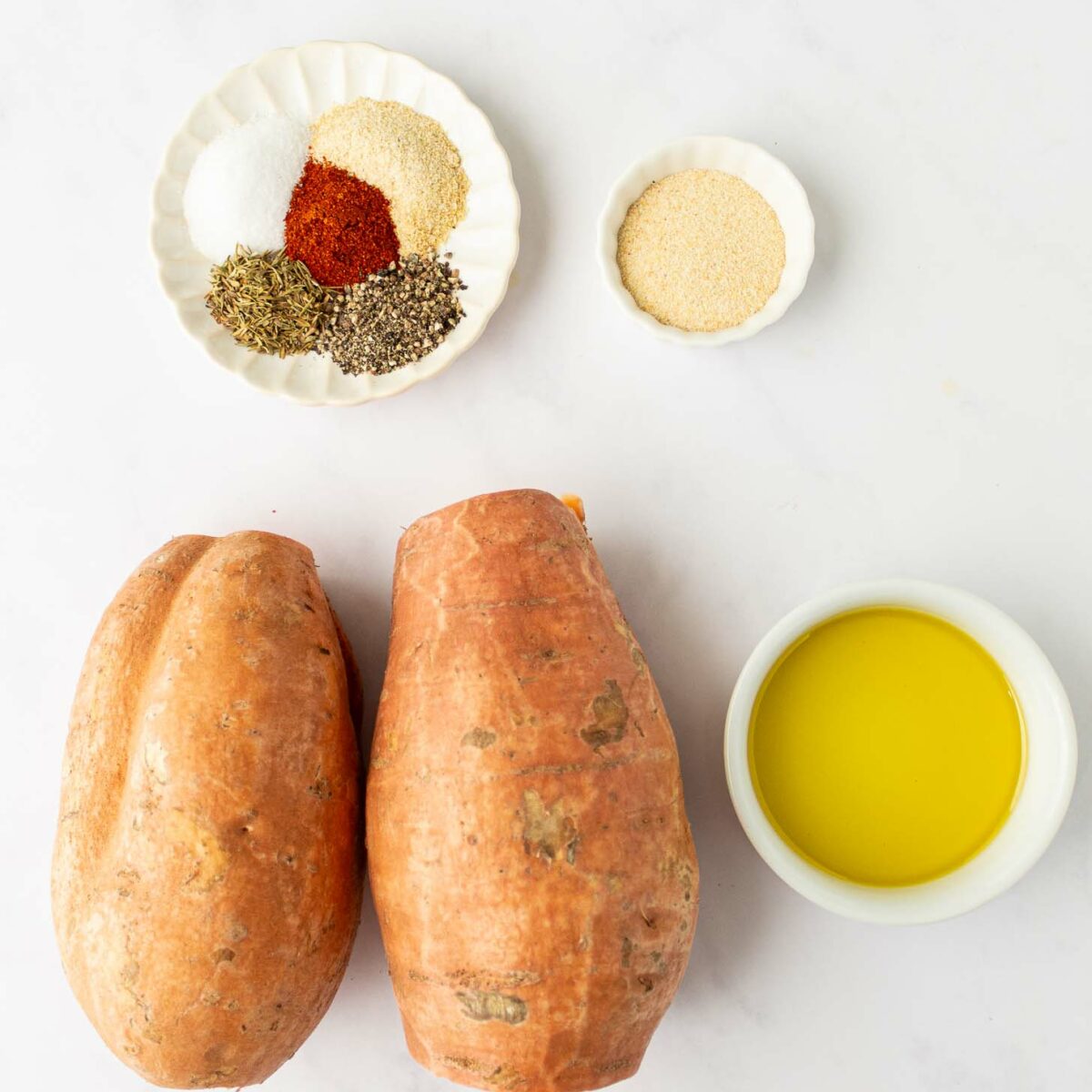 Two whole sweet potatoes, a small bowl of olive oil, a small dish with seasonings (salt, pepper, paprika, garlic powder, dried thyme), and a small bowl of breadcrumbs are arranged on a white surface.